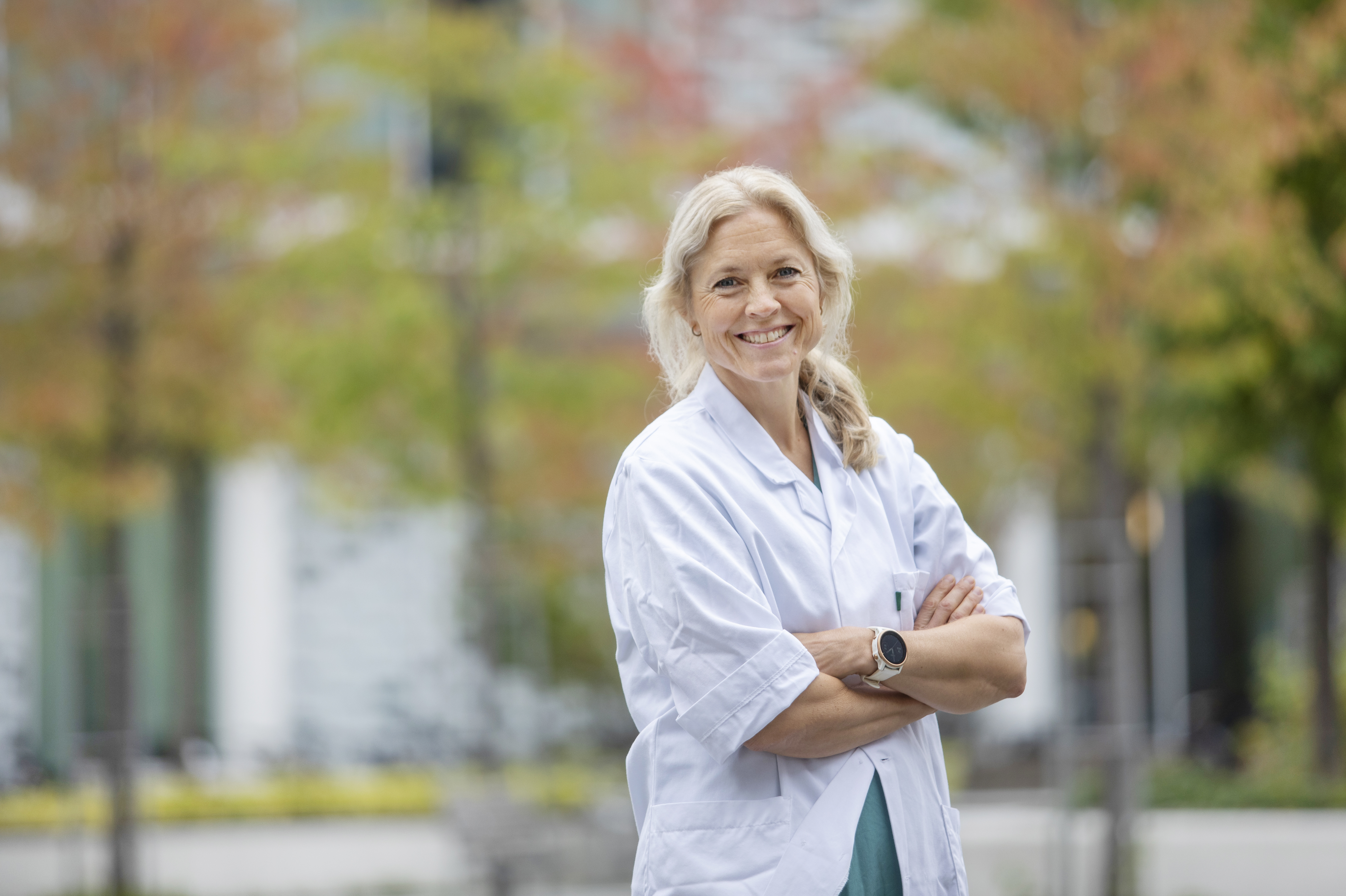 Smilende forsker i frakk står utenfor et sykehus med armene i kors