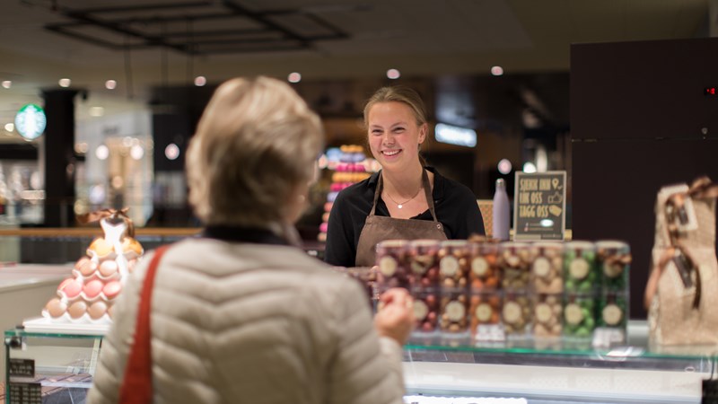 Bilde av en eldre dame som står ved en handledisk og en smilende ekspeditør hjelper henne.