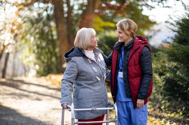 Bilde av en helsearbeider som smiler mot en eldre dame i rullator